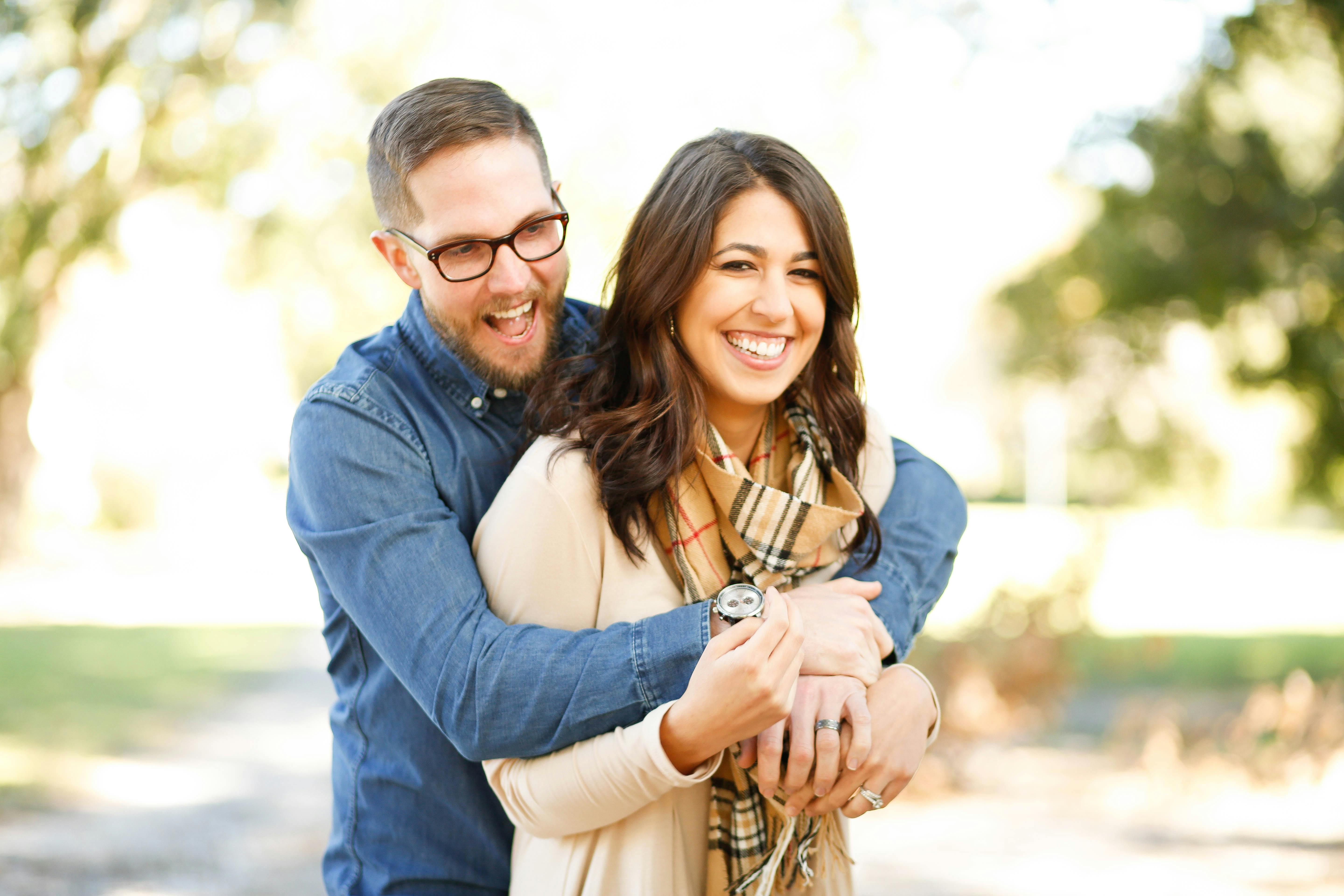 Young couple, man and woman, pose for a photo in the park. Man is wearing a denim shirt and eyeglasses, hugging the woman from behind. She has shoulder-length brown hair and is wearing a cream sweater and brown tartan scarf.