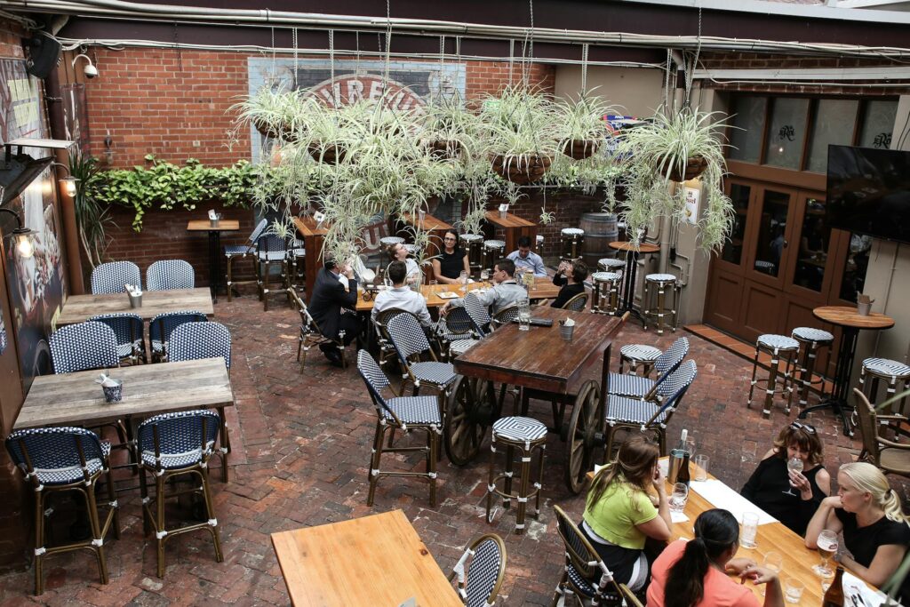 An outdoor restaurant with brick paver floor, hanging baskets of plants and quaint wooden tables set around. A small group of friends are enjoying a chat and drinks at a couple of the tables.