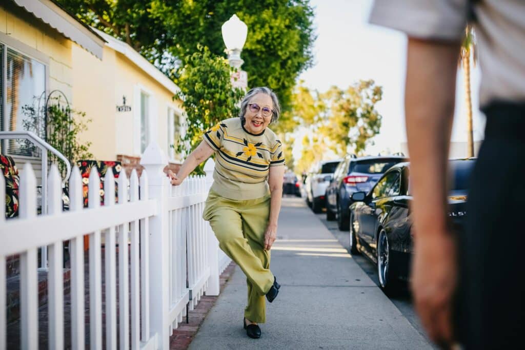 An older woman with grey hair in green pants and tshirt is leaning against a white picket fence to adjust her pantleg. She is smiling at the camera.