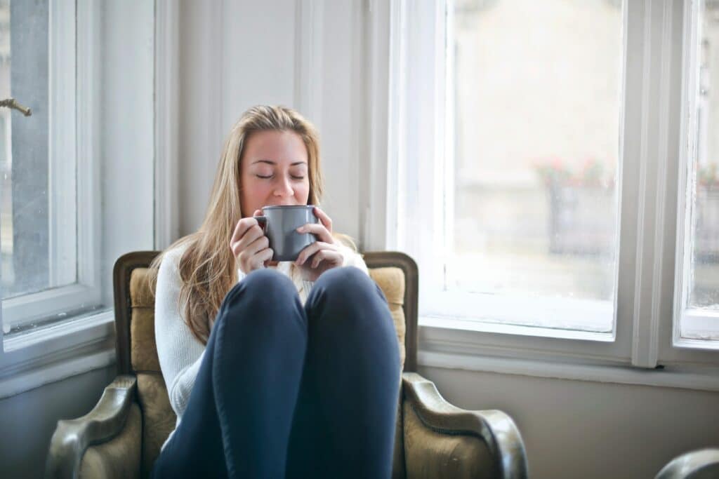 Blond woman sitting in an arm chair by big open windows in a white room holding a mug up to her face.