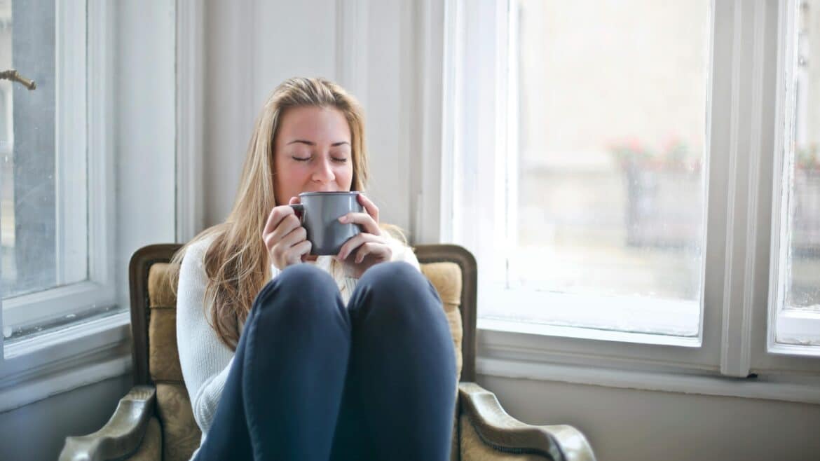 Blond woman sitting in an arm chair by big open windows in a white room holding a mug up to her face.