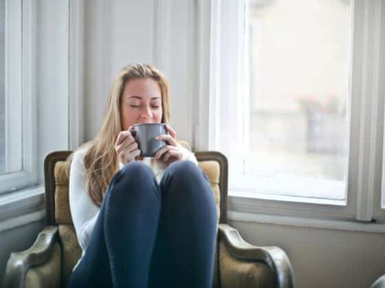 Blond woman sitting in an arm chair by big open windows in a white room holding a mug up to her face.