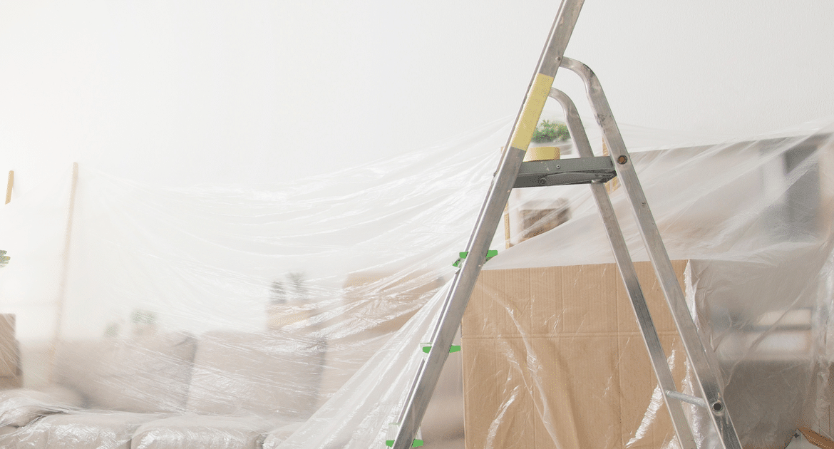 A living room mid-renovation with furniture under plastic dust sheets, taped moving boxes and an aluminium stepladder