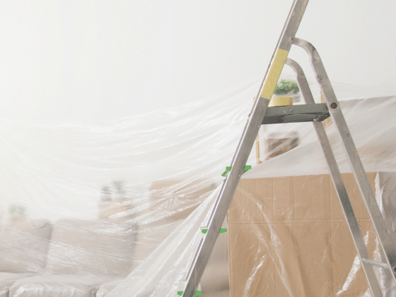 A living room mid-renovation with furniture under plastic dust sheets, taped moving boxes and an aluminium stepladder
